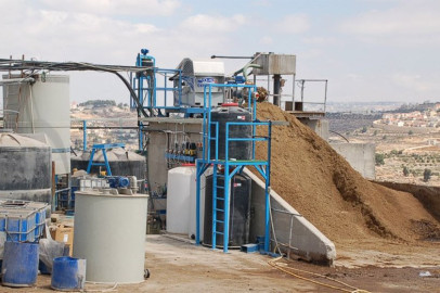 מתקן לטיפול במים עם מיכלים גליליים גדולים, צנרת כחולה ומכונות. תלולית גדולה של בוצה מעובדת נראית מימין. המתקן ממוקם בחוץ עם רקע נופי של גבעות, בניינים ושמיים מעוננים חלקית.