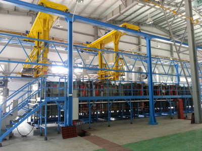 מתקן תעשייתי גדול הכולל סדרה של עגורנים עיליים צהובים על מבני פלדה כחולים. הסצנה כוללת פלטפורמות מתכת שונות, מעקות ומכונות תחת גג גבוה עם מספר אורות וחלונות. הרצפה היא שילוב של אריחים ירוקים ובז'.