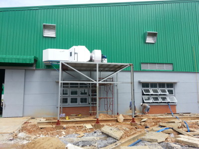 מבט חיצוני של בניין עם חלק עליון מתכת גלי ירוק וחלק תחתון אפור. שני עובדים מבצעים התאמות מתחת ליחידת מיזוג מוגבהת. הקרקע מכוסה בחומרי בנייה ופסולת.