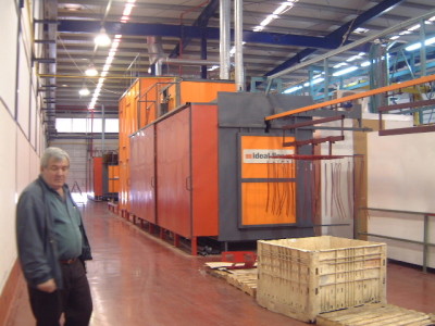 גבר לבוש ז'קט ירוק עומד במפעל ליד מכונה תעשייתית כתומה שכותרתה "אידיאל". למתקן תקרה גבוהה עם קורות חשופות, והרצפה בצבע חום-אדמדם. בחזית יש ארגז עץ גדול.