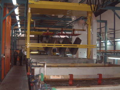 פנים מפעל עם עובדים המפעילים מכונות צהובות גדולות המשמשות לטבילת מבני מתכת במיכלי נוזל. המיכלים הם חלק מתהליך תעשייתי, אולי לציפוי או טיפול ברכיבי המתכת. צינורות ולוחות בקרה גלויים.