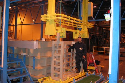 אדם עומד ליד מכונות תעשייתיות גדולות בתוך מפעל. נראה שהמכונות, הצבועות בצהוב ואפור, מרימות או מובילות רכיבי מתכת. האדם לובש תלבושת כהה ומביט לעבר המצלמה. בסביבת המפעל יש מבני מתכת וציוד שונים.