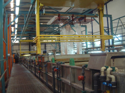 מבט בתוך מתקן תעשייתי, המציג פס ייצור מורכב עם מיכלי מתכת מרובים וצינורות ושסתומים בצבעים שונים. נראות מערכות עגורן עילי עם מסגרות צהובות, והאזור מואר היטב בתאורת פלורסנט.