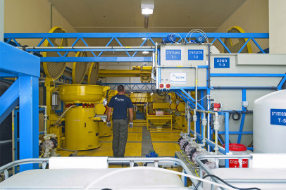 פועל עומד במתקן תעשייתי מואר עם מכונות שונות, צינורות ולוחות בקרה. הציוד ברובו כחול וצהוב, וניכרים שלטים עם טקסט. העובד לבוש בחולצה כהה ומכנסיים.