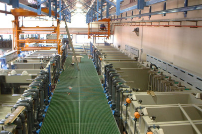 התמונה מציגה מתקן תעשייתי עם מיכלים גדולים ומערכות צנרת לאורך שני צידיו של שביל מתכת מרכזי. מעל, יש מנופים ומנגנוני הרמה, כשברקע פועל ליד התקרה. האזור נראה נקי ומאורגן היטב.