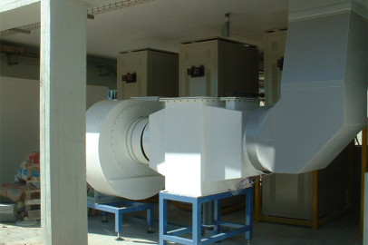 במתקן מותקנת מערכת אוורור תעשייתית גדולה עם מאוורר פליטה לבן ותעלות. הוא מורכב מכמה חלקי מתכת מחוברים המורכבים על מעמדים כחולים. לוחות בקרה חשמליים נראים ברקע.