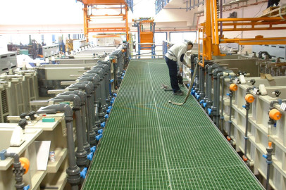 עובד בחולצה לבנה ומכנסיים כהים מפעיל ציוד במתקן תעשייתי. הסביבה כוללת צינורות רבים, שסתומים ומיכלים מלבניים גדולים. העובד עומד על שביל מגורר ירוק שעובר בין הציוד.