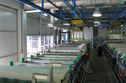 מתקן תעשייתי גדול עם מיכלי מתכת מלבניים רבים, מלאים בנוזל ומחוברים בצינורות וצינורות שונים. אדם לובש בגדי עבודה הולך לאורך שביל מגורר ליד הטנקים. מעל הראש, יש קורות תמיכה כחולות ומכונות.