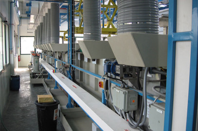 מתקן תעשייתי נקי ומודרני הכולל שורה ארוכה של מכונות וציוד מיוחדים. החדר מואר היטב ומאורגן עם תעלות, לוחות בקרה וצינורות המשולבים במערך. הרצפה עשויה בטון, ובחזית יש פח אשפה שחור.