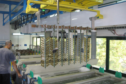 עובד מפעיל מכונות במפעל שנראה כאילו הוא מציף חלקי מתכת. ציוד עילי צהוב מחזיק חתיכות מתכת מעל מיכלי נוזל, כשברקע נראים צינורות, חוטים וצינורות. חלונות גדולים מספקים אור טבעי לחלל העבודה.